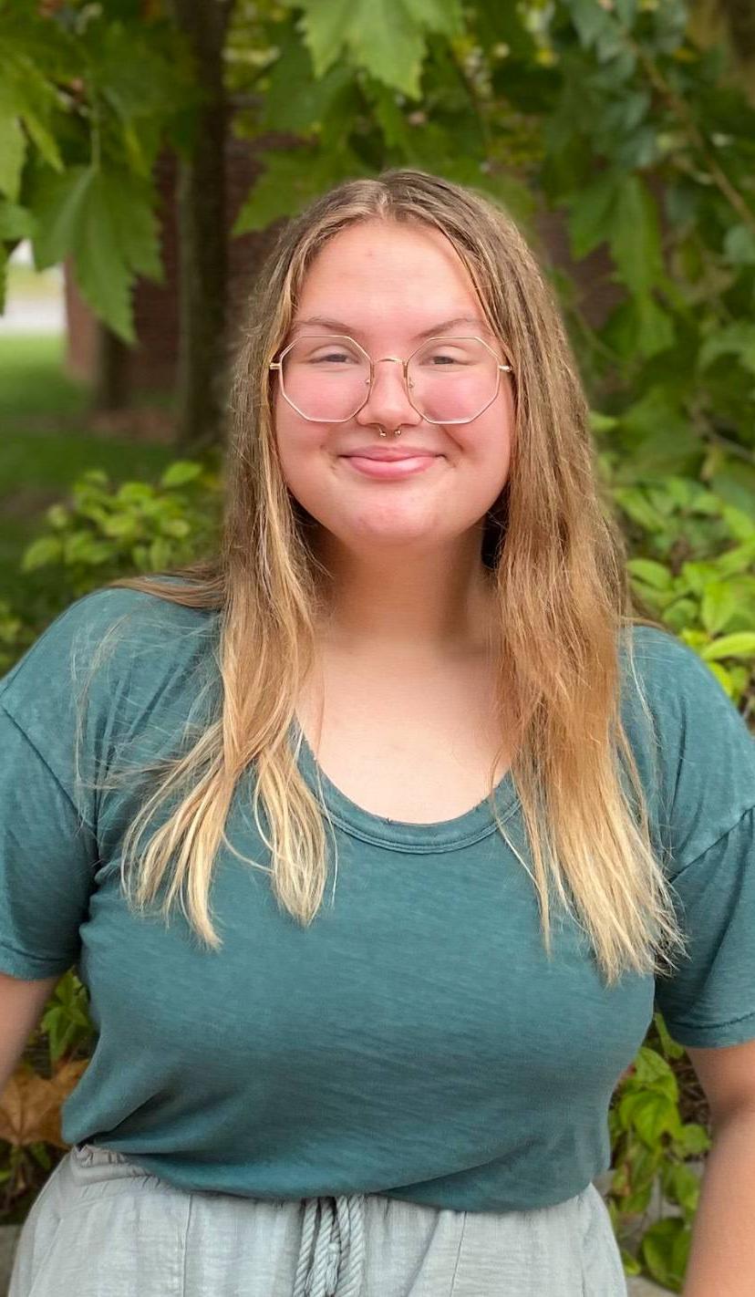 farren outside in a teal shirt and gold-rimmed glasses smiling infront of a short brick wall and a green tree